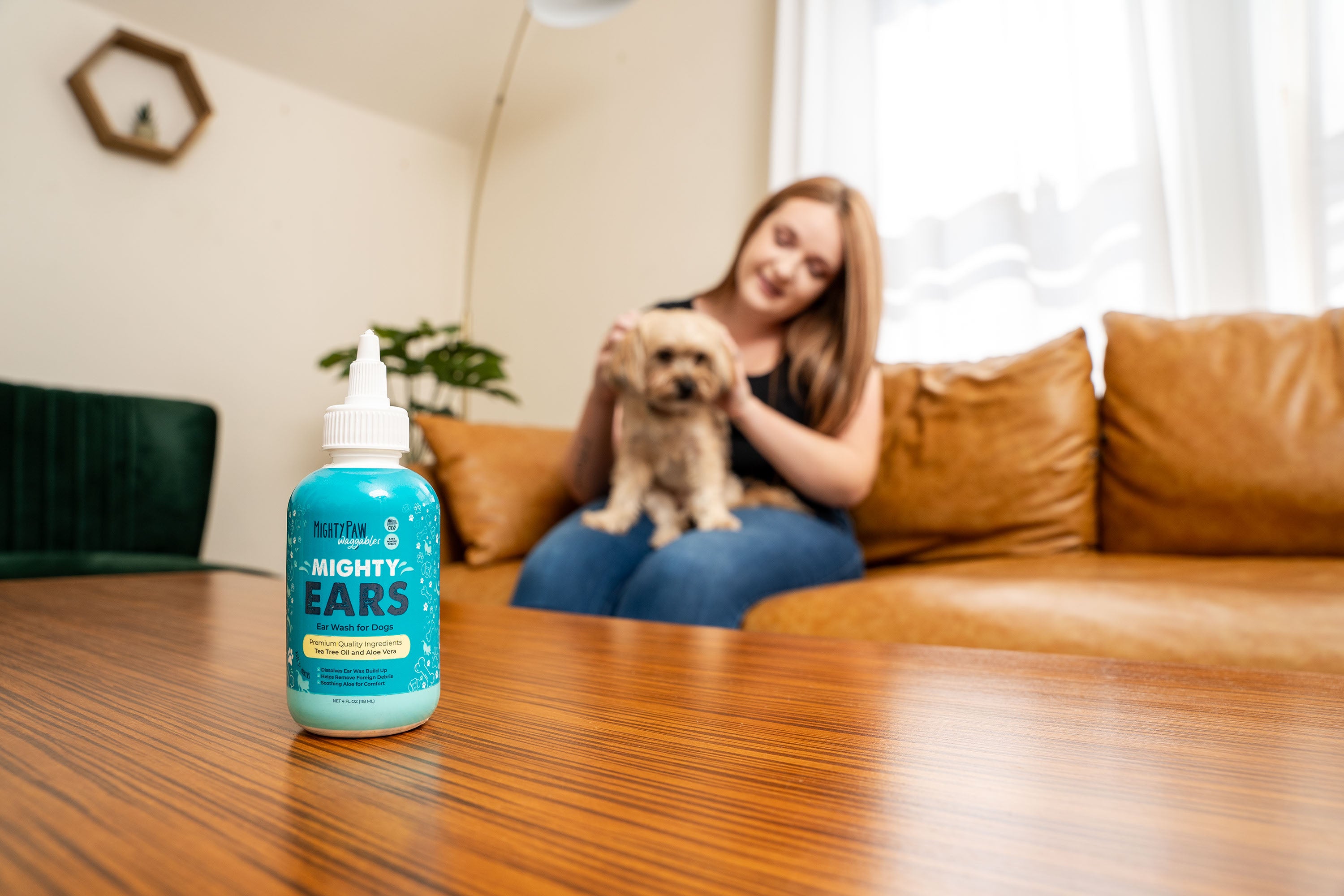 Woman sitting on couch petting her small tan dog with Mighty Ears ear wash on table in front of them.