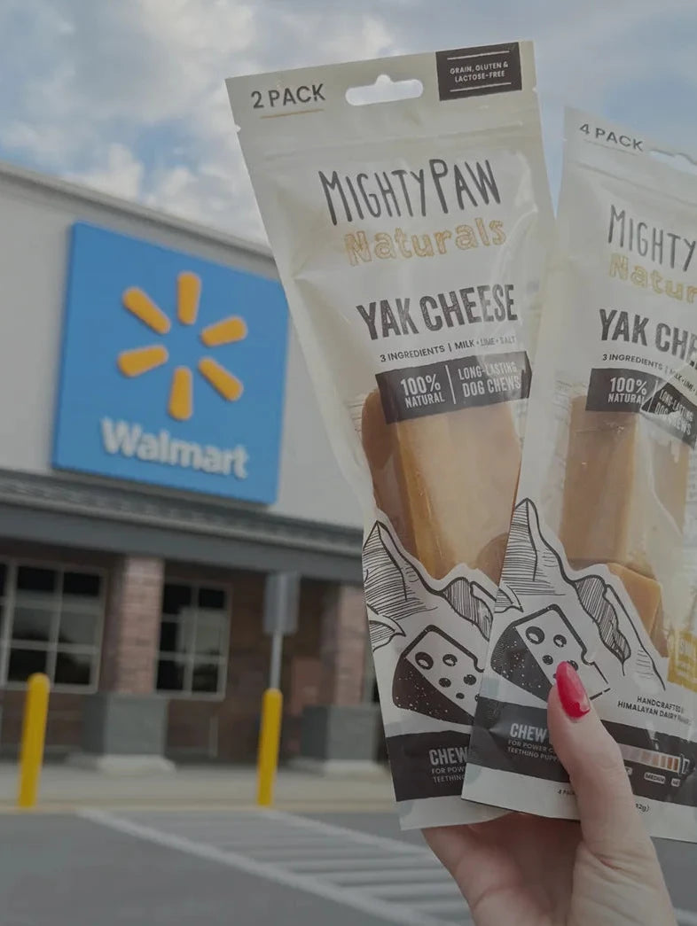 A hand with red nail polish holds two packages of Mighty Paw Naturals Yak Cheese dog treats in front of a Walmart store in the background.