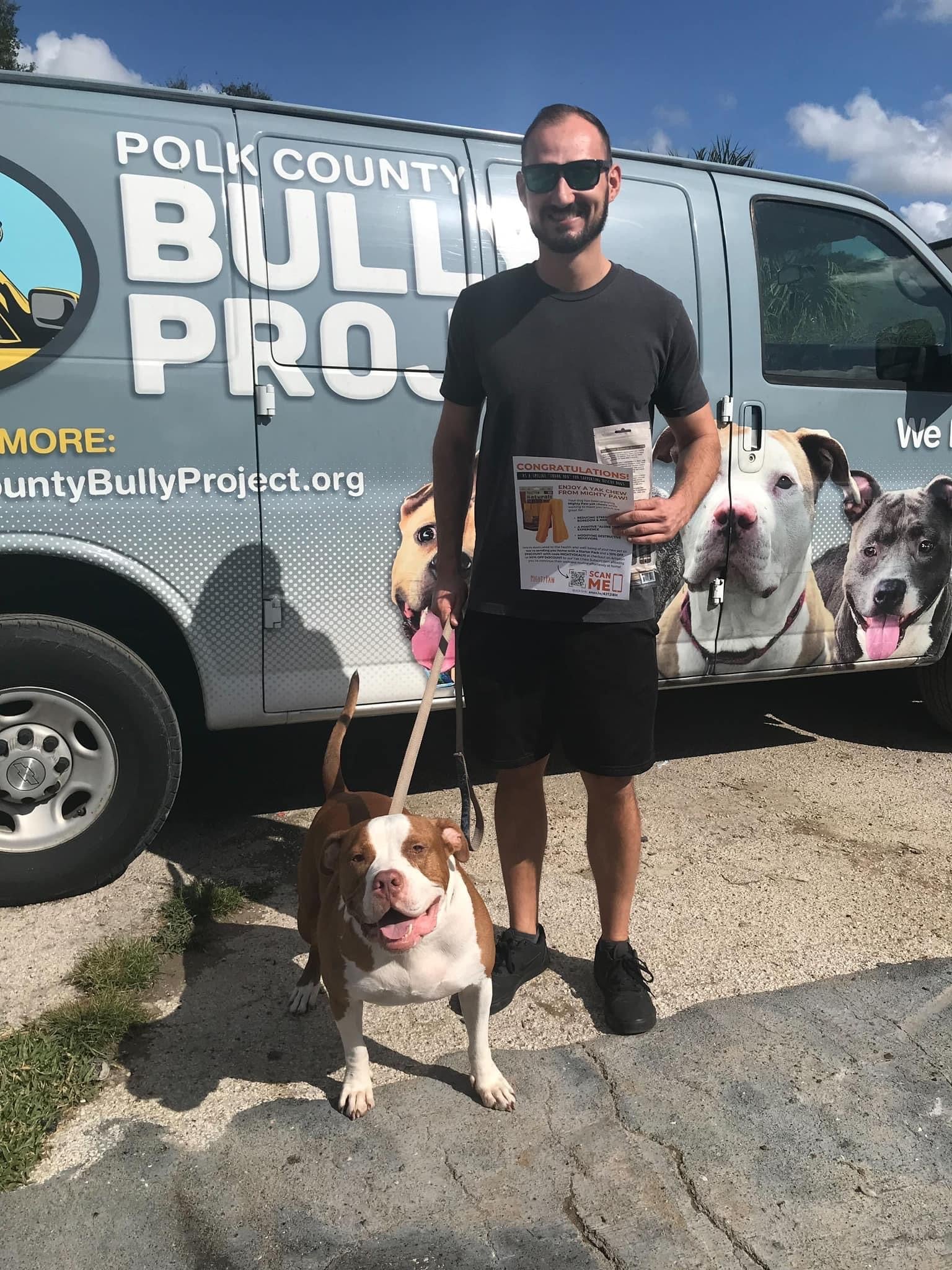 Man and his newly adopted brown and white dog stand in front of Polk County Bully Project van outside.