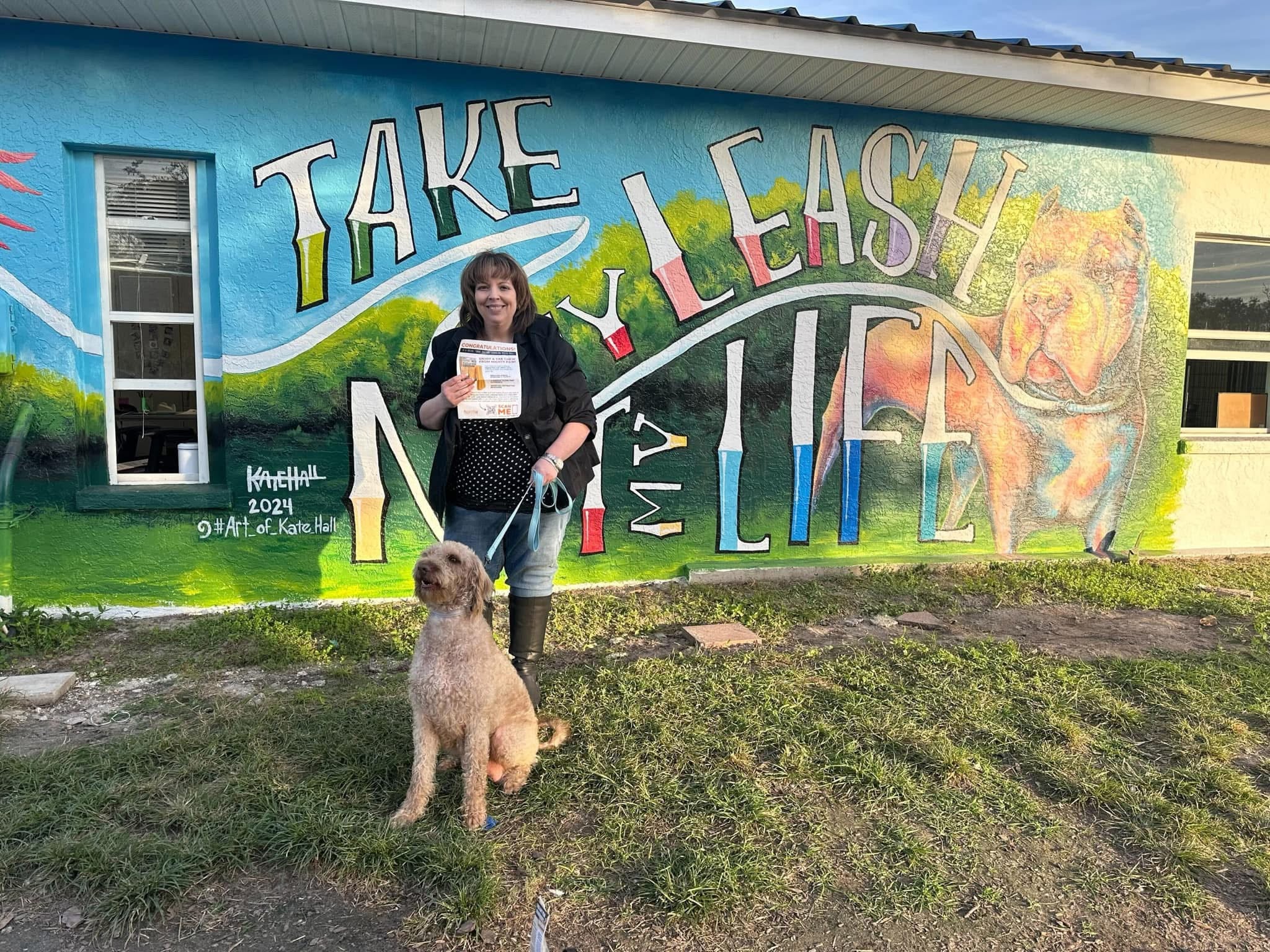 Woman with her dog stand outside in front of mural that says "Take My Leash Not My Life."