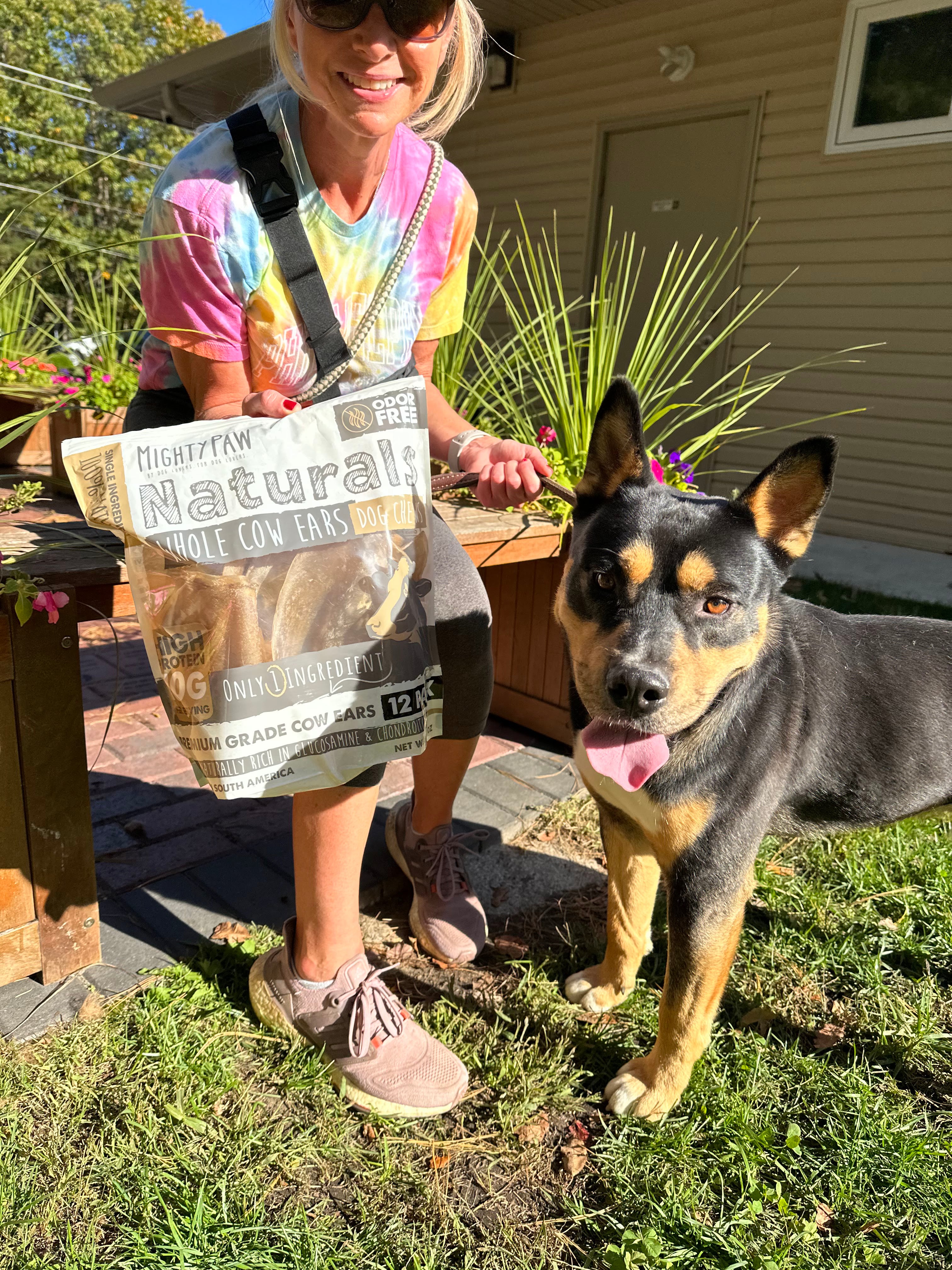 Woman with blonde hair holding Mighty Paw Cow Ears bag and black and brown dog sit outside.