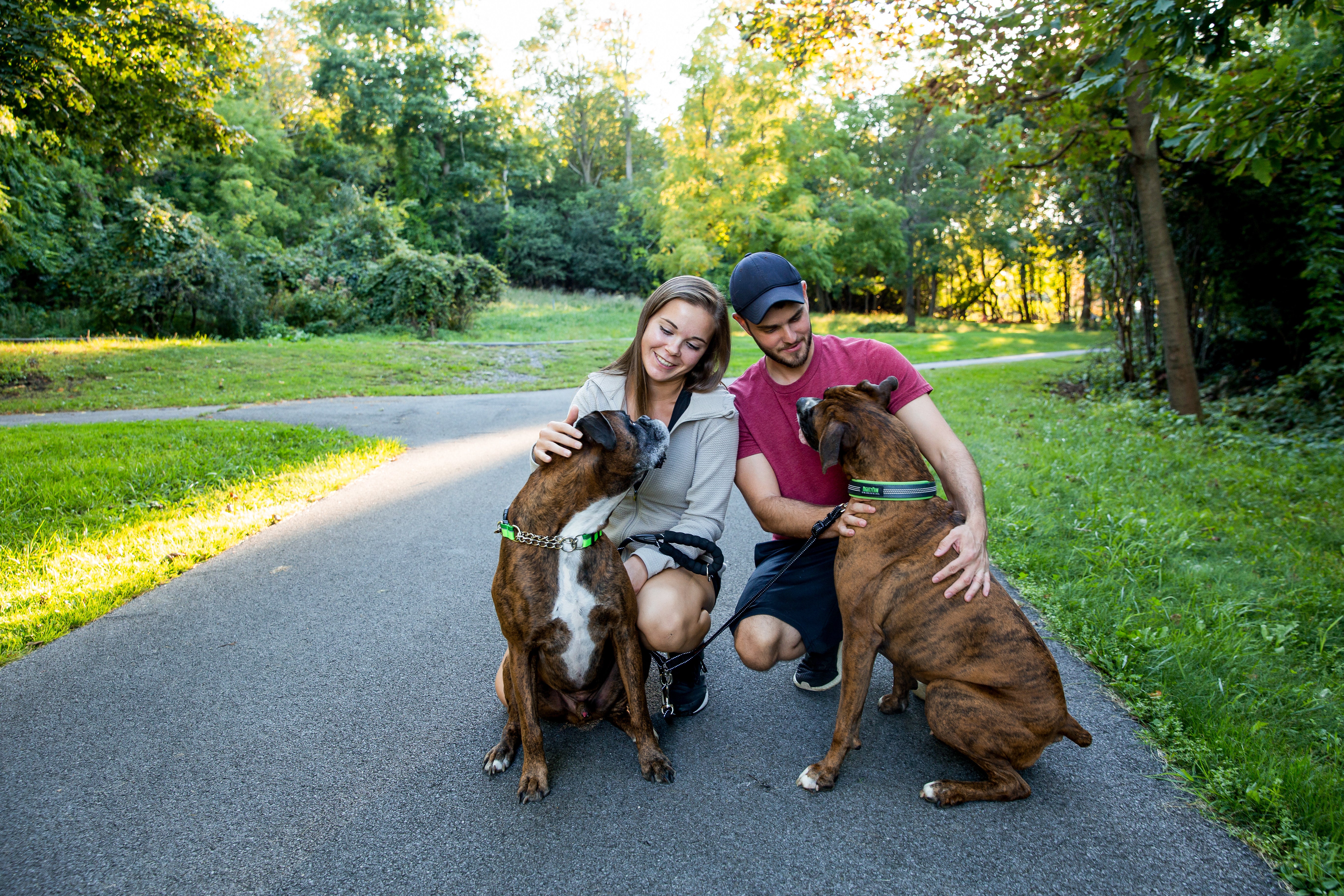 Man and woman sit outside in driveway with their two Boxer dogs.