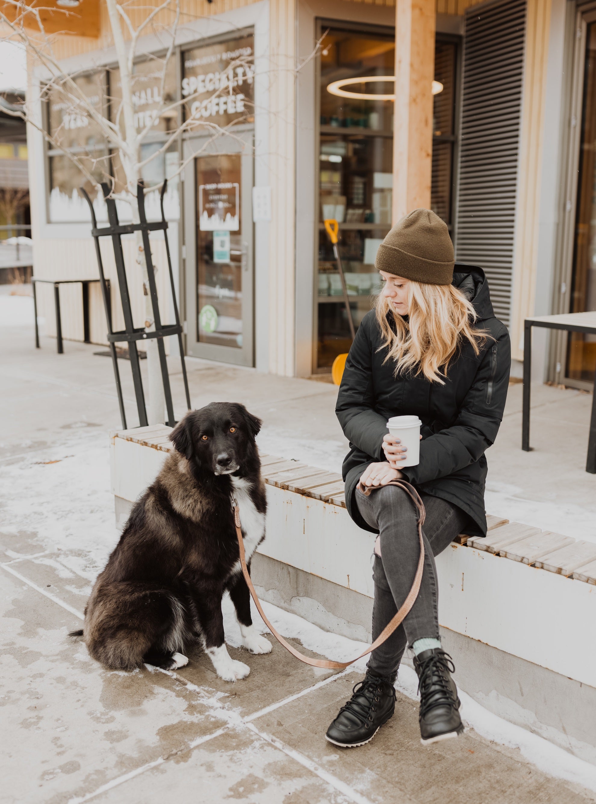 Blonde woman in black coat and pants sit outside coffee shop with big black dog on Mighty Paw Leash.